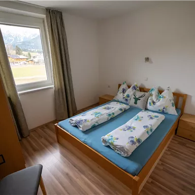 Ein einfaches Schlafzimmer mit einem Doppelbett und einem hellen Holzfußboden. Die Fenster lassen viel Tageslicht herein und die Wände sind hell gestrichen.