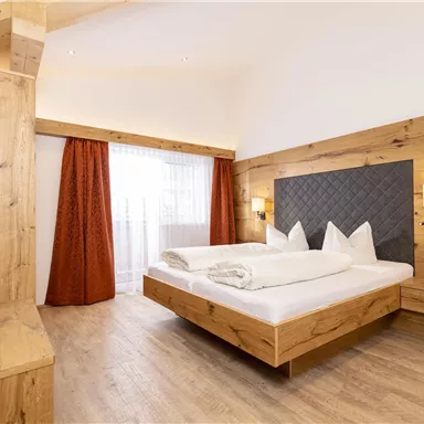 Ein modernes Schlafzimmer mit Holzverkleidung und einem großen Bett. Die Fenster lassen viel Tageslicht herein und die Vorhänge sind in warmen Orangetönen gehalten.