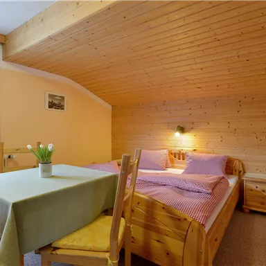 A cozy bedroom with a wooden ceiling and two beds. Next to the beds is a table with chairs and a flower vase.
