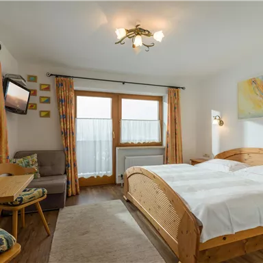 A cozy room with a double bed, a television, and a small table. The large window front provides plenty of daylight.