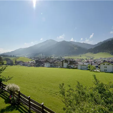 Eine malerische Landschaft mit grünen Wiesen und einem kleinen Dorf. Im Hintergrund sind hohe Berge und ein klarer blauer Himmel zu sehen.