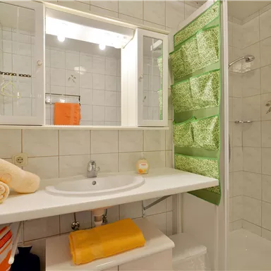 Ein modernes Badezimmer mit einer Dusche, einem Waschbecken und einem großen Spiegel. Handtücher in warmen Farben liegen ordentlich auf dem Regal.