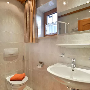 Ein modernes Badezimmer mit einer Dusche und einem Waschbecken. Es gibt helle Fliesen und orangefarbene Handtücher.