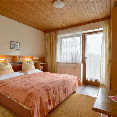 Ein gemütliches Schlafzimmer mit einem Doppelbett und warmen Holzdetails. Es gibt Fenster mit Vorhängen, die viel Licht hereinlassen.