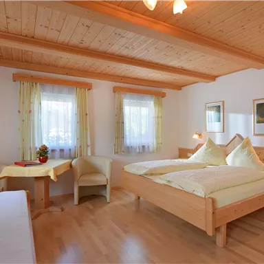 A cozy bedroom with a wooden ceiling and two beds. The room is bright and friendly with large windows and a small table.