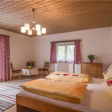 Ein gemütliches Schlafzimmer mit zwei Betten und bunten Kissen. Der Raum hat Holzdecke, Fenster mit Vorhängen und einen kleinen Tisch mit Stühlen.