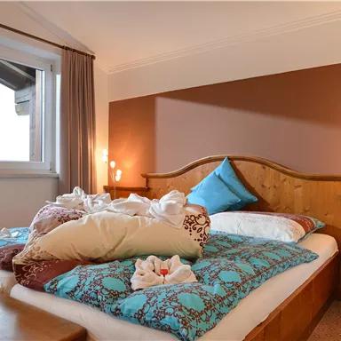 Ein gemütliches Schlafzimmer mit einem großen Bett und bunten Kissen. Die Fenster lassen viel Tageslicht herein und eine Orchidee steht auf der Fensterbank.