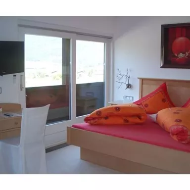Ein modernes Schlafzimmer mit einem großen Bett, bunten Kissen und einem Schreibtisch. Es gibt einen Fernseher und einen Aussichtsbalkon mit Blick auf die Landschaft.