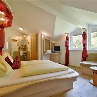 A cozy room with a modern design and large windows. There is a double bed, a seating area, and decorative curtains in red.