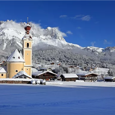Eine malerische Winterlandschaft mit schneebedeckten Hügeln und charmanten Hütten. Im Vordergrund steht eine gelbe Kirche vor einem klaren blauen Himmel.