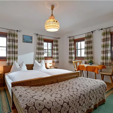 Ein gemütliches Schlafzimmer mit einem Doppelbett und rustikalen Möbeln. Die Fenster sind mit karierten Vorhängen dekoriert und es gibt eine kleine Sitzecke am Tisch.