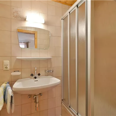 Ein modernes Badezimmer mit einer Dusche, einem Waschbecken und einem Spiegel. Die Wände sind hell gefliest und es gibt einige Handtücher.