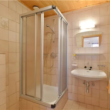 Ein Badezimmer mit einer Duschkabine, einem Waschbecken und einem Spiegel. Die Wände sind hell gefliest und die Decke ist aus Holz.