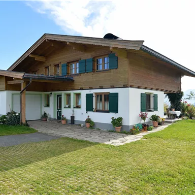 A beautiful two-story wooden house with a green lawn and blooming plants. The sky is clear and sunny.