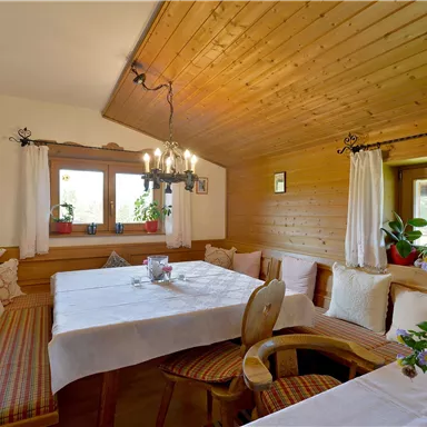 A cozy dining area with a wooden ceiling and bright furniture. Large windows let in plenty of light and plants decorate the space.