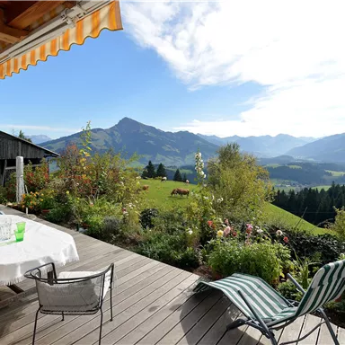 A terrace with a table and chairs, surrounded by flowers. In the background, green hills and mountains stretch under a blue sky.