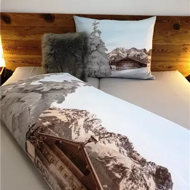 Ein gemütliches Bett mit einer Bettdecke und Kissen im Bergdesign. Im Hintergrund sind hölzerne Elemente und sanftes Licht zu sehen.