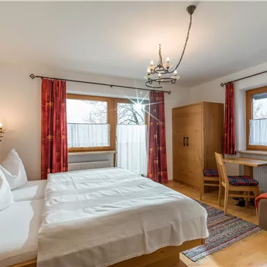 A cozy bedroom with two beds, wooden surfaces, and warm colors. Large windows let in plenty of light and offer a view outside.