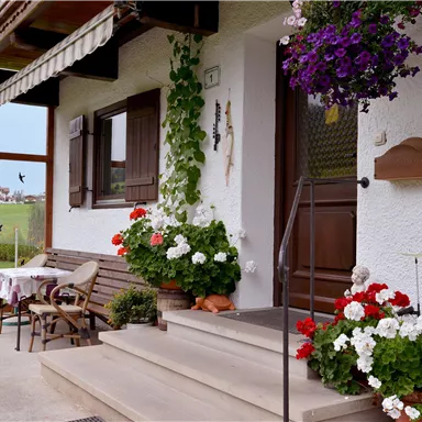 Ein einladender Eingangsbereich mit blühenden Pflanzen und einem Tisch. Die Fassade des Hauses ist weiß, mit Holzverzierungen und einem schönen Ausblick auf die Landschaft.