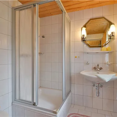 Ein modernes Badezimmer mit einer Duschkabine und einem weißen Waschbecken. Die Wände sind hell gefliest und es gibt einen großen Spiegel.