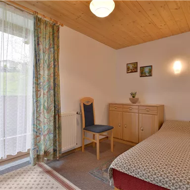 Ein gemütliches Zimmer mit einem Einzelbett und einem Stuhl. Es gibt Fenster mit Vorhängen und eine Holzdecke, die eine warme Atmosphäre schafft.