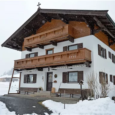 Ein traditionelles Alpenhaus mit Balkonen, umgeben von schneebedecktem Gelände. Der Himmel ist bewölkt, und es fällt leichten Schnee.
