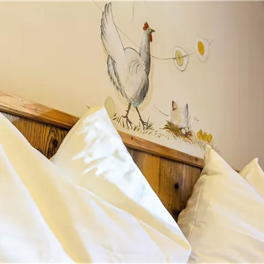 Ein gemütliches Schlafzimmer mit zwei großen, weißen Kissen und einer Hühnermalerei an der Wand. Die Dekoration sorgt für eine entspannte, ländliche Atmosphäre.