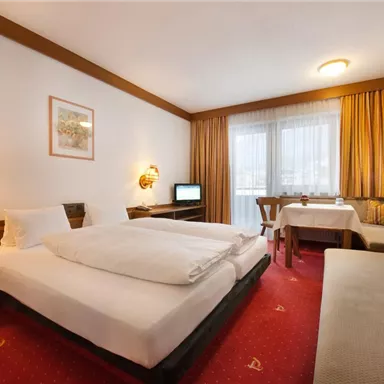 Ein helles Hotelzimmer mit zwei Betten, einem Tisch und einem gemütlichen Sitzbereich. Große Fenster sorgen für viel Tageslicht.