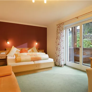 Ein gemütliches Schlafzimmer mit einem großen Bett und bunten Kissen. Es gibt einen Balkon mit Aussicht ins Freie und eine angenehme, helle Atmosphäre.