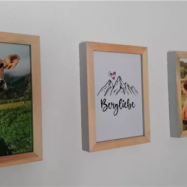 Drei Bilder hängen an einer Wand. Zwei zeigen Kühe, und eines hat den Schriftzug „Bergliebe“ mit einer Bergillustration.