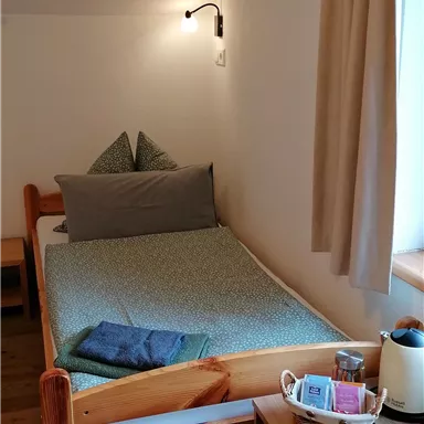 Ein einfaches Schlafzimmer mit einem Einzelbett, Kissen und Decke. Auf einem Tisch befinden sich ein Becher und einige persönliche Gegenstände.