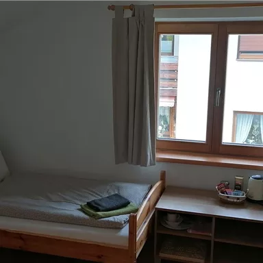 Ein einfaches Zimmer mit einem Einzelbett und einem Tisch. Es gibt ein Fenster mit Vorhängen, das Tageslicht hereinlässt.