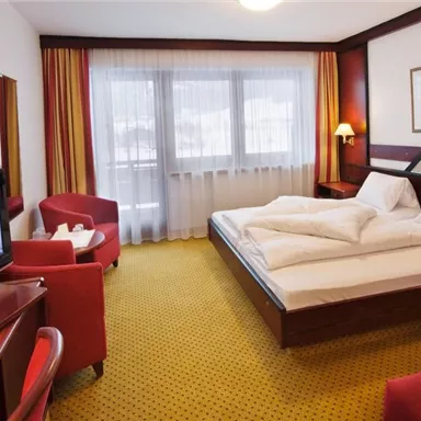 A comfortable hotel room with a large double bed and a television. The decor is modern and inviting, with a bright carpet and large windows.
