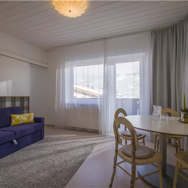 Ein modernes Wohnzimmer mit einem blauen Sofa und einem runden Esstisch. Große Fenster lassen viel Licht herein und verleihen dem Raum ein einladendes Ambiente.