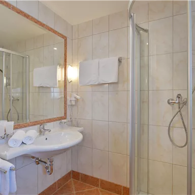 Ein modernes Badezimmer mit einer Dusche, einem Waschbecken und einem großen Spiegel. Handtücher sind ordentlich aufbewahrt und die Wände sind in hellen Tönen gefliest.