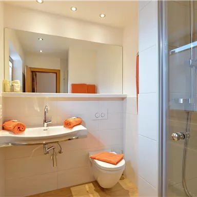 Ein modernes Badezimmer mit hellen Fliesen und einem großen Spiegel. Auf dem Waschbecken liegen orangefarbene Handtücher, und es gibt eine komfortable Dusche.