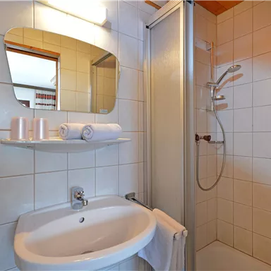 Ein modernes Badezimmer mit einer Dusche und einem Waschbecken. Die Wände sind gefliest und es gibt Handtücher sowie Toilettenartikel auf dem Regal.