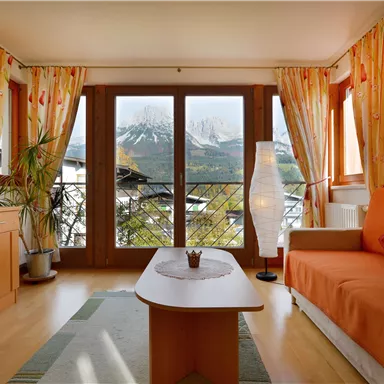 Ein gemütliches Wohnzimmer mit einem orangen Sofa und einem kleinen Tisch. Große Fenster bieten einen Blick auf die Berge und lassen viel Licht herein.