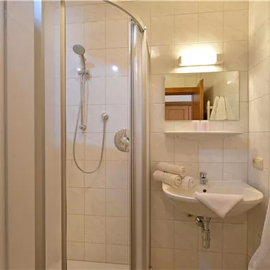 Ein modernes Badezimmer mit einer Duschkabine und einem Waschbecken. Weiße Fliesen und Handtücher verleihen dem Raum eine saubere und helle Atmosphäre.