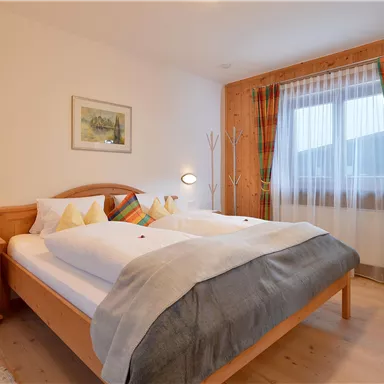 Ein gemütliches Schlafzimmer mit einem Doppelbett und Holzmöbeln. Große Fenster mit bunten Vorhängen sorgen für viel Licht.