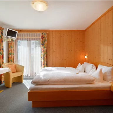 Ein gemütliches Zimmer mit Holzverkleidung, einem Doppelbett und zwei Stühlen. Große Fenster sorgen für viel natürliches Licht.