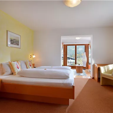 Ein helles Zimmer mit zwei Betten, warmen Farben und großen Fenstern. Es gibt gemütliche Möbel und viel Tageslicht.