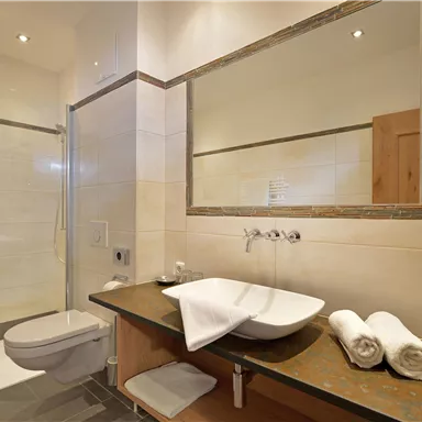 Ein modernes Badezimmer mit Dusche und einem großen Spiegel. Auf der Ablage befinden sich Handtücher und Pflegeprodukte.