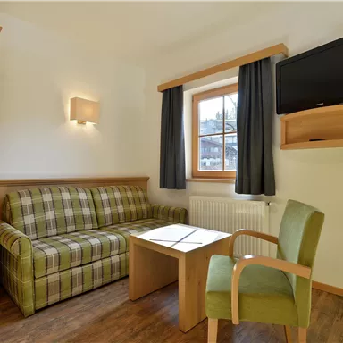 Ein gemütliches Wohnzimmer mit einem beigen Sofa und einem Tisch. An der Wand hängt ein Fernseher, und es gibt ein Fenster mit Vorhängen.