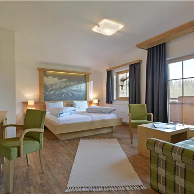 Ein helles Hotelzimmer mit einem großen Bett, gemütlichen Möbeln und Fenstern, die viel Licht hereinlassen. Die Einrichtung ist modern und einladend.