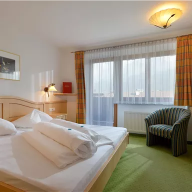 Ein gemütliches Hotelzimmer mit einem Doppelbett und frischen Handtüchern. Es gibt einen Sessel, einen Schreibtisch und große Fenster, die viel Licht hereinlassen.