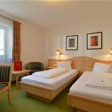 Ein einfaches Zimmer mit zwei Einzelbetten, einem Tisch und einem Stuhl. Die Wände sind hell und es gibt Fenster mit Vorhängen.