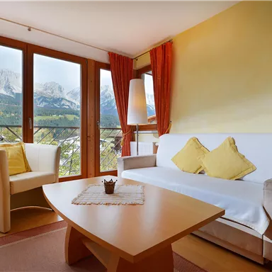 Ein gemütliches Wohnzimmer mit einem weißen Sofa und zwei gelben Kissen. Durch die Fenster sieht man die Berge im Hintergrund.