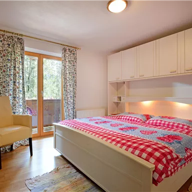 Ein helles Schlafzimmer mit einem großen Bett und bunten Bettbezügen. Es gibt einen Sessel und ein Fenster mit Blick ins Freie.