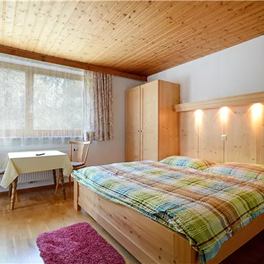 Ein gemütliches Schlafzimmer mit einem Doppelbett und einer Holzwand. Es gibt einen Tisch mit Stühlen und große Fenster, die viel Licht hereinlassen.